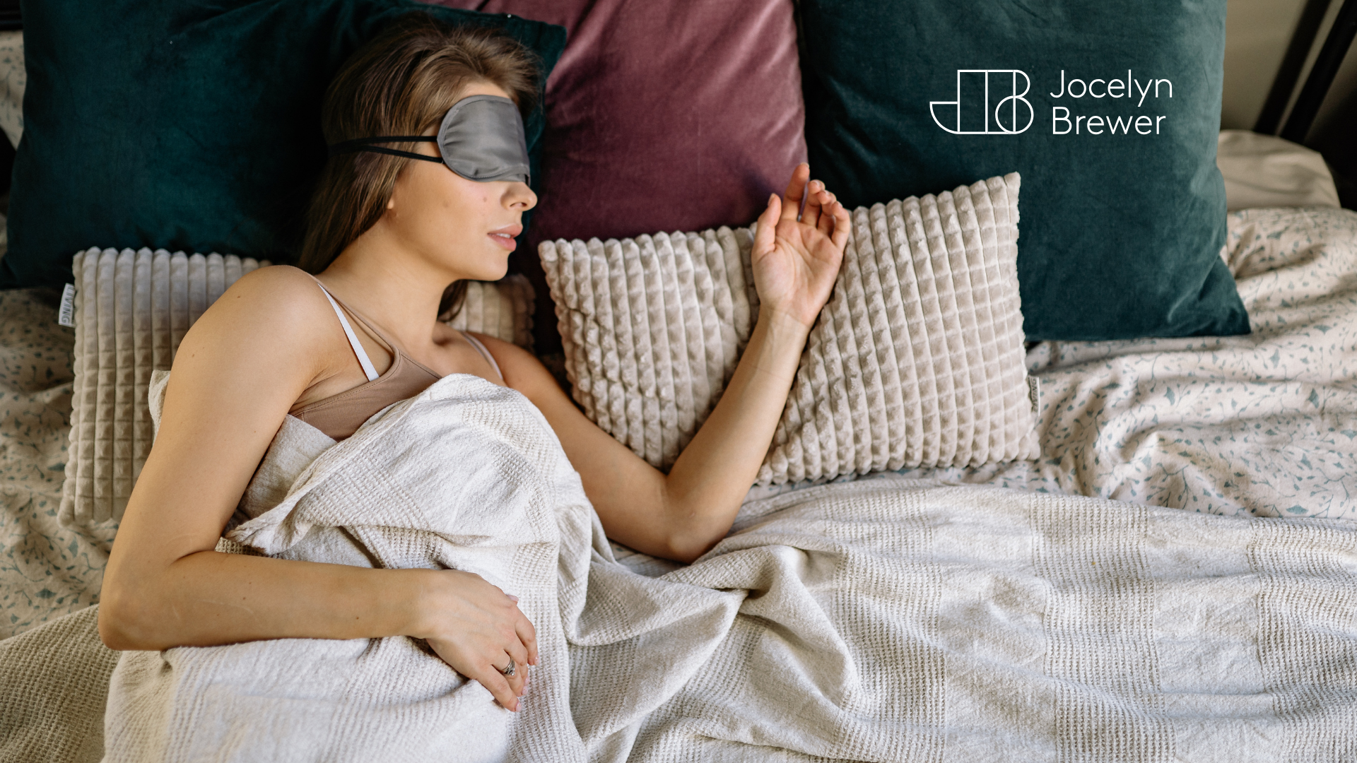 Woman sleeping peacefully in bed wearing a silk sleep mask, surrounded by soft pillows and neutral bedding, with Jocelyn Brewer branding in the corner.